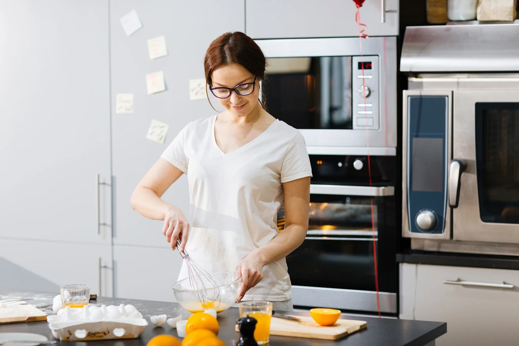 Katherine "Katie" Hayes, wearing a white t-shirt and glasses, focuses on whisking ingredients in a glass bowl in her kitchen.