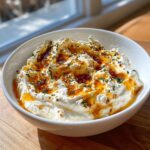 A white bowl filled with fluffy ricotta dip, drizzled generously with golden chili oil and sprinkled with fresh green herbs.