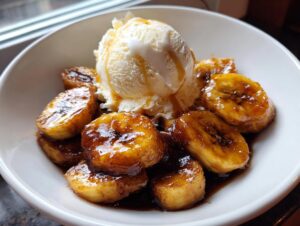 A white bowl filled with caramelized bananas in rich sauce, topped with a scoop of vanilla ice cream, ready to eat Bananas Foster.