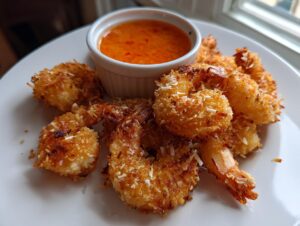 A plate of golden-brown, crispy coconut shrimp served with a small ramekin of bright orange sweet chili dipping sauce.