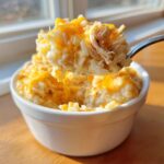 A spoonful of creamy, cheesy chicken and rice topped with melted cheddar cheese being lifted from a white bowl.