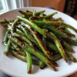 A close-up of bright green, blistered chinese green beans with garlic sauce served on a white plate.