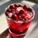 Close-up of a bright red cranberry pomegranate margarita garnished with whole cranberries, pomegranate seeds, and a sugar rim.