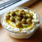 A close-up of creamy dirty martini dip garnished with whole green olives, olive oil, and herbs.
