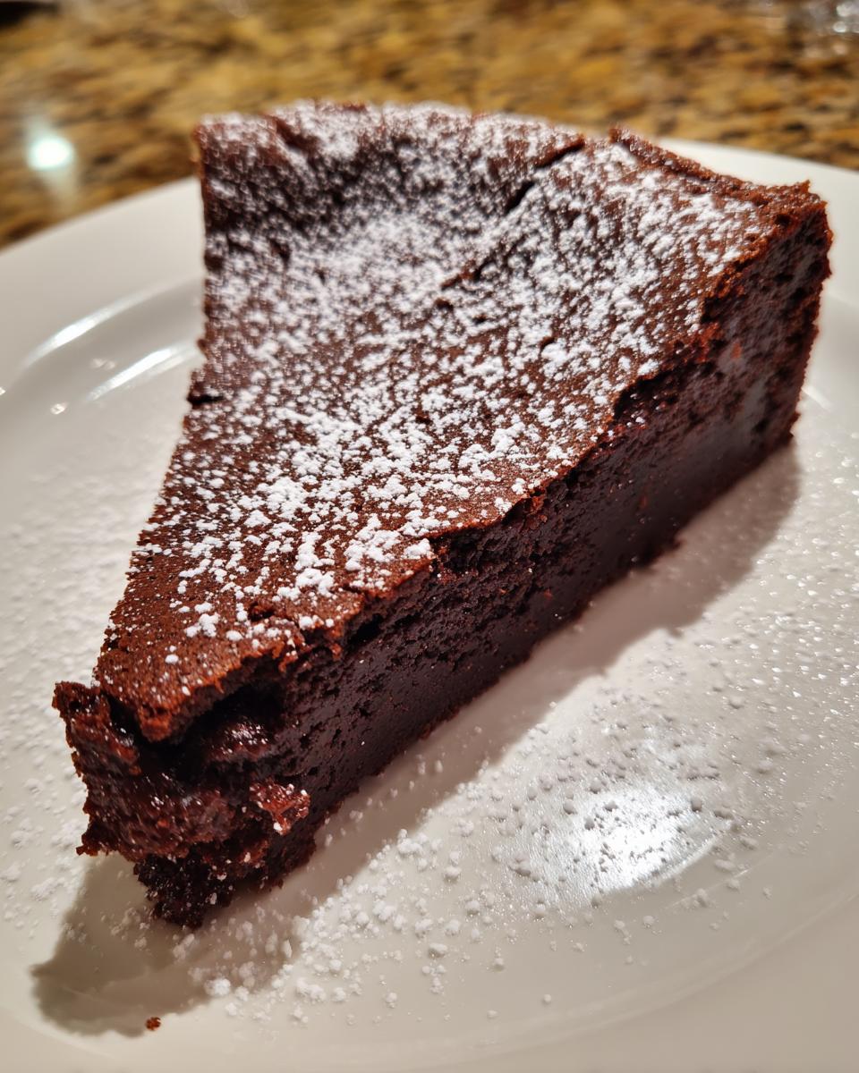 A rich, dark slice of flourless chocolate torte dusted generously with white powdered sugar on a white plate.