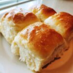 Four fluffy, golden-brown dinner rolls brushed with a shiny glaze, resting on a white plate.