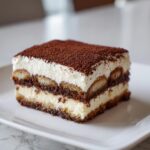 A square slice of creamy Tiramisu layered with coffee-soaked ladyfingers and dusted heavily with cocoa powder.