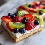 Close-up of a square slice of fruit pizza sugar cookie topped with cream cheese frosting, strawberries, blueberries, and kiwi.