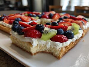 A close-up side view of a slice of fruit pizza sugar cookie topped with cream cheese frosting and fresh berries.