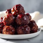 A close-up of a pile of rich, dark Grape Jelly Meatballs glistening with a thick, sweet glaze on a white serving dish.