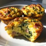 Three golden-brown egg muffin cups, one with a bite taken out, showing ham and spinach filling.