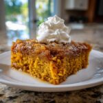 A moist slice of pumpkin dump cake topped with a dollop of whipped cream, served on a white plate.