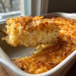 A spoonful of creamy, cheesy hashbrown casserole being lifted from a white baking dish, showing a crispy topping.
