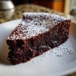 A decadent slice of moist flourless chocolate torte dusted generously with white powdered sugar on a white plate.
