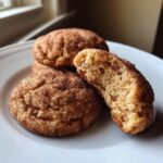 Close-up of three soft Snickerdoodle cookies on a white plate, one broken open showing a chewy interior.