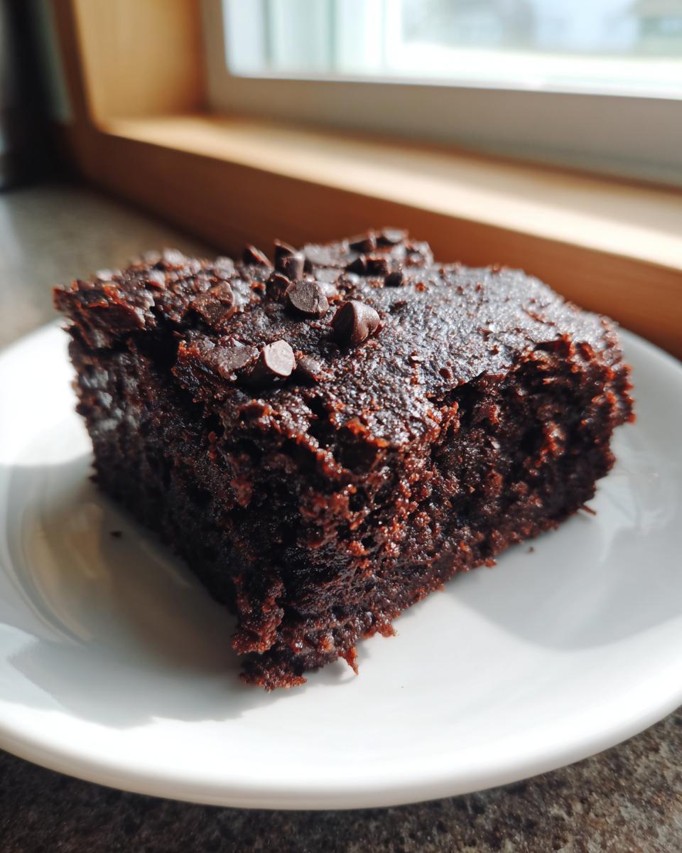 A close-up of a rich, dark chocolate square of sweet potato brownies topped with chocolate chips, sitting on a white plate.