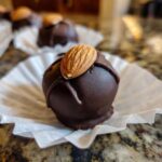 A decadent, dark chocolate coated almond joy truffles resting in a white paper liner, topped with a whole almond.