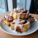 A stack of four fluffy, freshly baked cinnamon roll treats generously covered in white cream cheese icing.