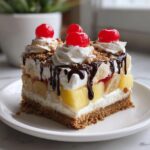 A decadent slice of banana split cake featuring layers of fruit, cream, and a graham cracker crust, topped with whipped cream and cherries.