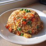 A mound of colorful cauliflower fried rice mixed with scrambled egg, bright orange carrots, and green peas served on a white plate.