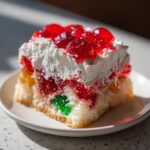 Close-up of a moist slice of christmas poke cake featuring layers of colorful jello and whipped cream topping.