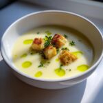 A bowl of creamy white garlic soup topped with golden croutons, fresh thyme, and olive oil drops.