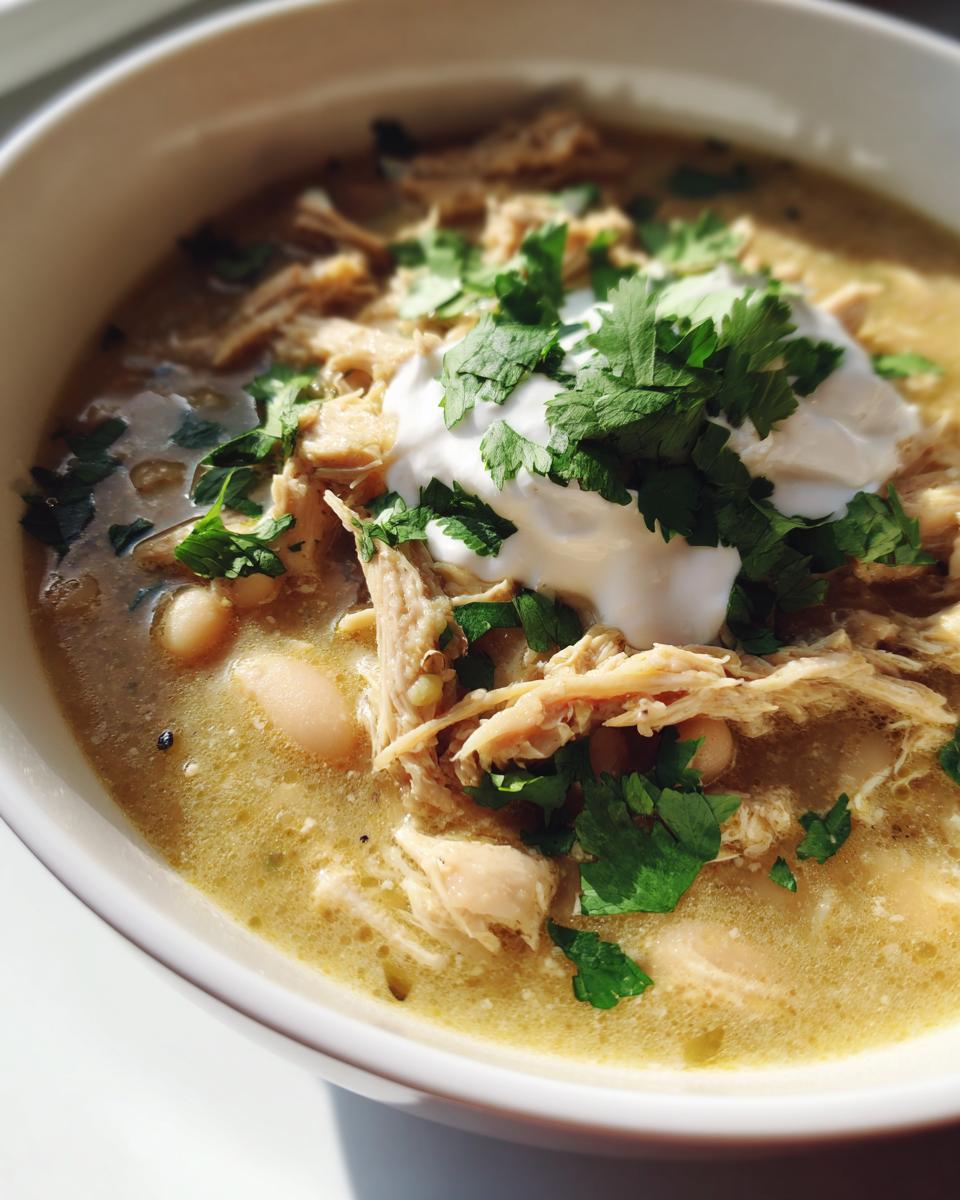 Close-up of a bowl of crockpot white chicken chili topped with shredded chicken, sour cream, and fresh cilantro.