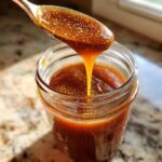 A spoonful of thick, glossy salted caramel sauce drips back into a glass jar filled with the rich caramel.