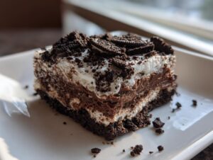 A rich, square slice of 4-layer dirt cake featuring chocolate pudding, cream, and crushed Oreos on a white plate.