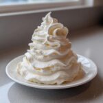 A tall, perfectly piped swirl of fluffy vanilla buttercream frosting sitting on a small white plate.