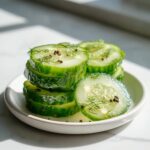 Stack of bright green cucumber slices seasoned for refrigerator pickles, topped with fresh dill and peppercorns.