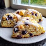 Four flaky lemon blueberry scones topped with white glaze served on a white plate near a window.
