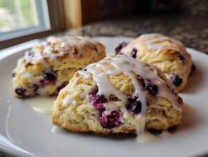 Three triangular lemon blueberry scones drizzled with thick white glaze, sitting on a white plate near a window.