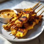 Close-up of grilled, golden yellow chicken satay skewers served on a plate next to a small bowl of peanut sauce.