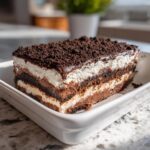 A close-up of a decadent slice of 4-layer dirt cake showing alternating layers of chocolate cake, cream, and pudding, topped with chocolate frosting and cookie crumbs.