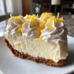 A close-up of a slice of creamy lemon pie with a graham cracker crust, topped with piped whipped cream and bright lemon zest.
