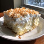 A close-up of a moist slice of coconut poke cake topped with whipped cream and toasted coconut flakes.