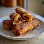 Close-up of crispy, golden peach cobbler egg rolls dusted with cinnamon sugar, one cut open showing the filling.