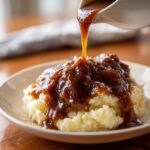 Close-up of rich homemade brown gravy being poured from a gravy boat onto a mound of fluffy mashed potatoes.
