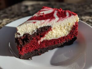 A decadent slice of red velvet cheesecake featuring layers of dark crust, bright red cake, creamy white cheesecake, and a marbled topping.