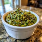 Close-up of vibrant, chunky roasted green tomato salsa with visible red pepper flakes in a small white bowl.
