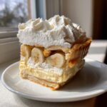 A thick slice of banana cream pie showing layers of crust, custard, banana slices, and topped with high peaks of whipped cream.