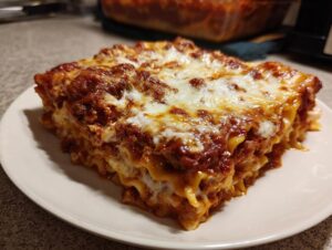 A thick, square slice of baked ravioli layered with meat sauce and melted cheese, served on a white plate.