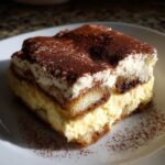 A rich, layered slice of homemade tiramisu recipe, dusted heavily with cocoa powder, sitting on a white plate.