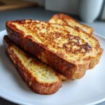 Close-up of three thick slices of golden brown sourdough french toast stacked on a white plate.