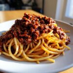 Close-up of spaghetti topped generously with rich, dark red bolognese sauce on a white plate.