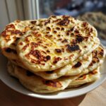 A stack of golden, fluffy homemade naan bread with dark, charred bubbles, glistening slightly with butter or ghee.