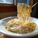 A fork lifts fettuccine noodles coated in rich, creamy homemade alfredo sauce, showing extreme cheese stretch.