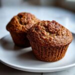 Two perfectly baked, dark brown bran muffins sitting on a white plate.