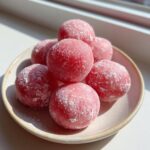A stack of vibrant pink watermelon mochi dusted with white powder, resting on a small ceramic plate.